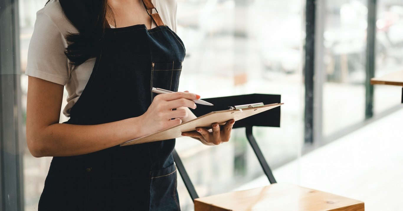 Persona con delantal negro tomando notas en una libreta en un ambiente luminoso.